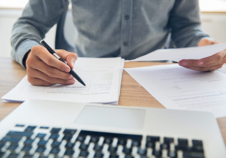 Unrecognizable businessman sitting at table holding ballpoint pen and papers and checking documents in office
