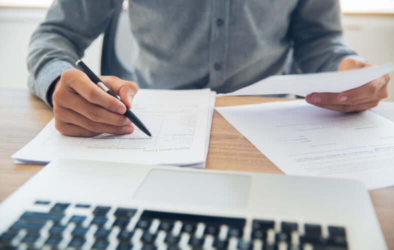Unrecognizable businessman sitting at table holding ballpoint pen and papers and checking documents in office