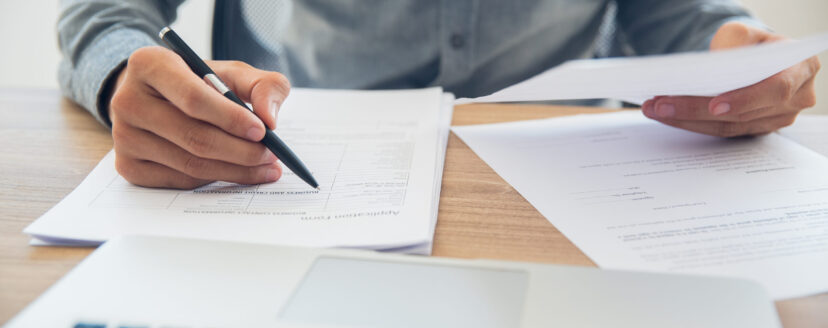 Unrecognizable businessman sitting at table holding ballpoint pen and papers and checking documents in office
